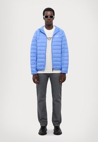 Jachetă puffer de culoare bleu deschis, cu design matlasat, logo negru, tricou alb cu grafică, blugi închiși la culoare și pantofi negri, purtată cu ochelari de soare.
