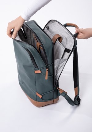 Dark green leather backpack with tan accents, dual zippers, and a grid-patterned interior. Features two compartments and a padded strap.