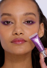 Tube cosmétique violet tenue près du visage, présentant une finition brillante. Le modèle porte un fard à paupières violet et une couleur de lèvres douce. L'arrière-plan est neutre.