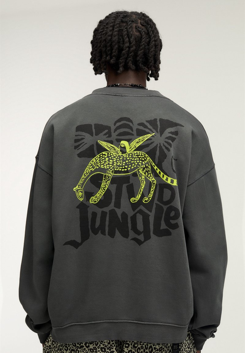 Personne aux cheveux tressés portant un sweat-shirt sombre avec un animal ailé jaune néon et un texte noir "Studio Jungle" dans le dos.
