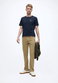 Maglietta blu navy a girocollo, pantaloni slim-fit kaki, sneakers bianche, che tiene una felpa verde oliva; design minimalista e toni neutri.