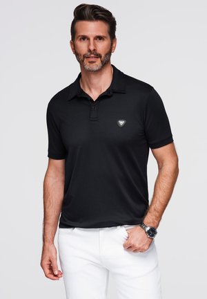Man met een baard, gekleed in een zwart polo-shirt met korte mouwen en witte broek, staat met één hand in zijn zak tegen een witte achtergrond.