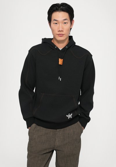 Black hoodie with contrasting orange stitching, adjustable drawstring, kangaroo pocket, and leather toggle detail. Paired with brown striped pants.