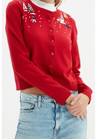 Cardigan rosso ritagliato con texture a coste, caratterizzato da bottoni e motivi ricamati a tema invernale, indossato sopra un dolcevita bianco.