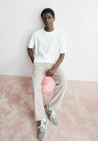 Wit katoenen T-shirt, lichtgrijze broek en grijze sneakers met witte accenten. Onderwerp zittend op een roze kruk tegen een lichte achtergrond.