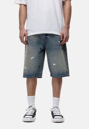 Shorts en denim bleu clair avec un délavage, présentant des détails déchirés et une coupe droite. Portés avec un t-shirt blanc et des baskets noires.