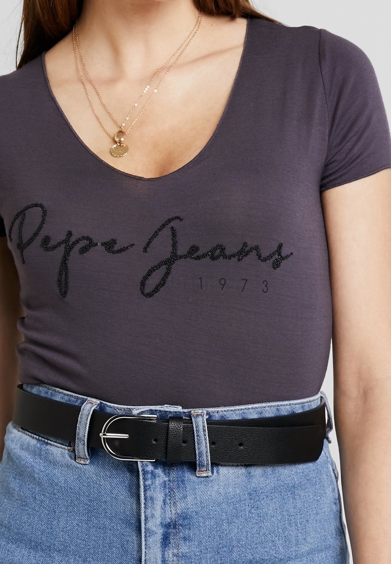 Šedé tričko s výstřihem do V, na kterém jsou flitrové nápisy "Pepe Jeans" a rok "1973", doplněné vysokými džínovými šortkami a černým koženým opaskem.
