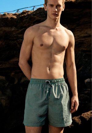 Jeune homme debout torse nu en short de bain vert, avec des falaises rocheuses et un ciel bleu en arrière-plan, en plein air sous le soleil.