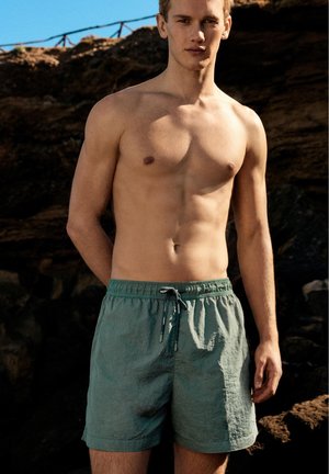 Ung mand står barbrystet i grønne badeshorts med klippeklædte skrænter og blå himmel i baggrunden, udendørs i sollys.