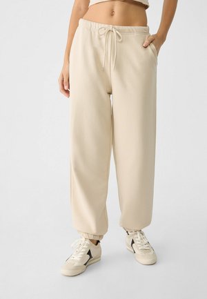 Femme portant un jogging beige à cordon de serrage et des baskets crème avec des accents noirs, debout devant un fond clair uni.