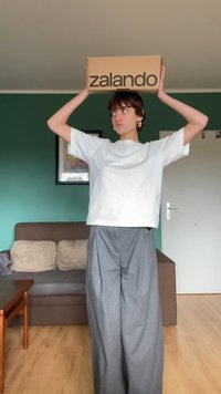 Barna Zalando doboz a személy feje fölött. Laid-back, fehér pólót és szürke, bő szárú nadrágot visel egy belső térben.