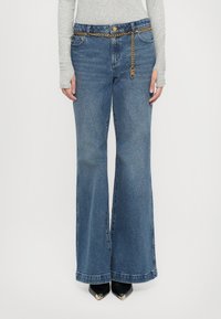 Personne portant un jean évasé bleu taille haute, une ceinture fine chaîne dorée, un haut gris à manches longues et des chaussures noires pointues avec des bouts dorés.