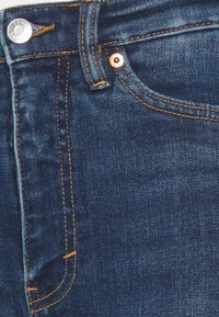 Primer plano de unos jeans de mezclilla azul oscuro que muestra el bolsillo delantero, la trabilla, los remaches de metal y los detalles de costura contrastante en naranja.
