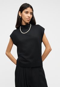 Femeie cu păr lung şi întunecat, purtând un top negru fără mâneci, un colier de perle şi pantaloni negri plisati, stând cu mâinile la spate.