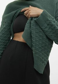 Zielony kardigan dziany o fakturze z motywem diamentowym, noszony na czarnej bluzce. Konstrukcja zawiera ściągacze przy mankietach i odsłonięty przód.