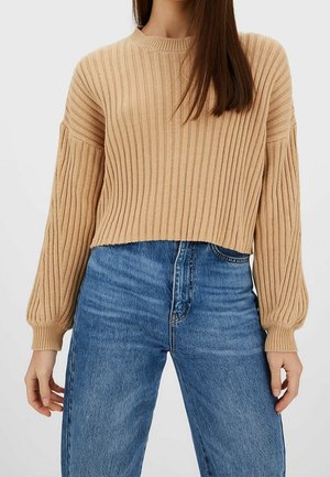 Kobieta nosząca beżowy sweter w prążki z bufkami na rękawach i niebieskie jeansy z wysokim stanem, pokazana od szyi do bioder.