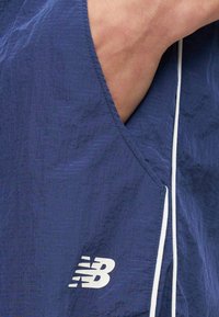 Χέρι στην τσέπη σκούρων μπλε σορτς New Balance με λευκά κορδόνια, που δείχνουν υφασμάτινη υφή και λογότυπο κοντά στο τελείωμα.