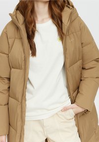 Chaqueta acolchada marrón con capucha, con paneles cosidos y cremallera frontal. Se lleva sobre una camiseta de cuello redondo de color claro y pantalones crema.