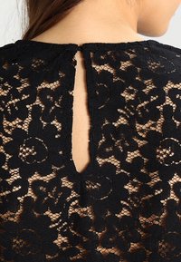 Bakvyn av en svart blommig spets-topp med ett nyckelhål vid halsringningen, som visar hud genom spetsmönstret.