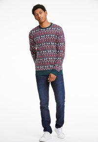 Sarkans un zils svītrots džemperis ar ziemeļbriežu un ģeometriskām apdrukām, ar apaļu apkakli un ribotiem piedurknēm, saskaņots ar tumšiem džinsiem.