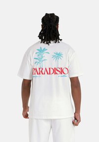Witte katoenen t-shirt met blauwe palmbomenafbeeldingen en rode tekst "PARADISIO" op de achterkant. Relaxed fit met korte mouwen.
