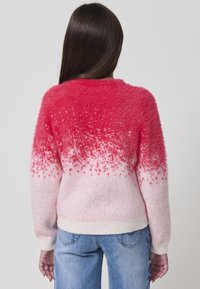 Femme aux longs cheveux noirs portant un pull flou dégradé rose-blanc et un jean bleu, se tenant face à un fond gris clair uni.