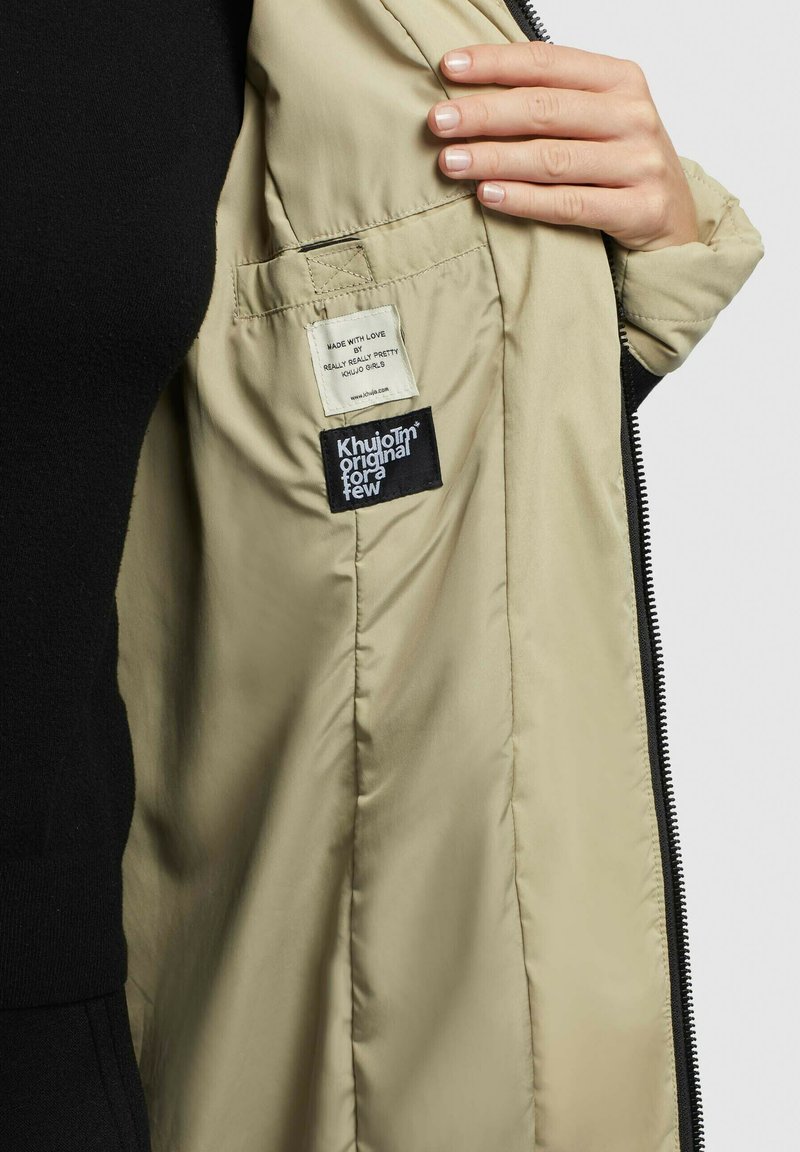 Beige gesteppte Jacke mit schwarzem Reißverschluss. Die Etiketten tragen die Aufschrift "Mit Liebe hergestellt von Really Really Pretty" und "Khujo™ original für eine kleine Auswahl."