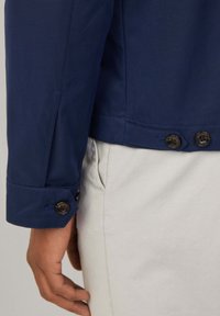 Chaqueta azul marino con un diseño estructurado, que presenta un puño abotonado y una textura suave, combinada con pantalones de color claro.