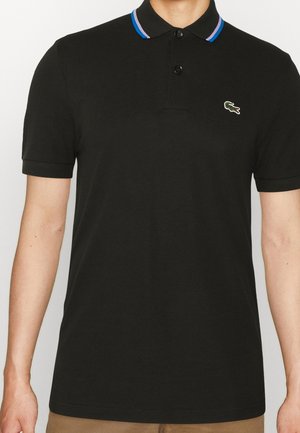 Hombre que lleva una camiseta polo negra de manga corta con una tapeta de dos botones y un borde del cuello a rayas azul y púrpura.