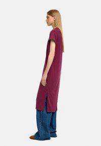 Burgundowa, oversize sukienka t-shirtowa z krótkimi rękawami i bocznymi szczelinami, stworzona z miękkiego materiału. Noszona z szerokimi niebieskimi spodniami dżinsowymi.