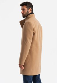Abrigo beige hasta la rodilla en suave mezcla de lana, con un cuello alto y un diseño minimalista, combinado con jeans oscuros.
