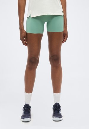 Άτομο που φοράει ανοιχτοπράσινα εφαρμοστά σορτς, λευκές κάλτσες crew, σκούρα μπλε αθλητικά παπούτσια και λευκό τοπ, σε απλό ανοιχτόχρωμο φόντο.