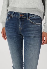 Jeans de mezclilla azul con una cintura de altura media, cinco bolsillos, un ligero desgaste y detalles de costura. Combinados con un sudadera gris claro.