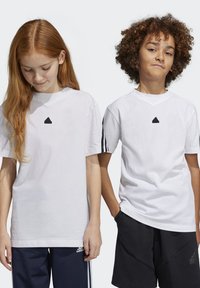 Dos niños llevan camisetas de algodón blancas. La camiseta de la niña tiene un logo negro; la camiseta del niño tiene ribetes negros en las mangas, combinada con pantalones cortos oscuros.