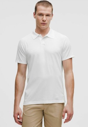 Młody mężczyzna ubrany w gładką białą koszulkę polo z krótkim rękawem i beżowe spodnie, stojący na tle jednolitego szarego tła.