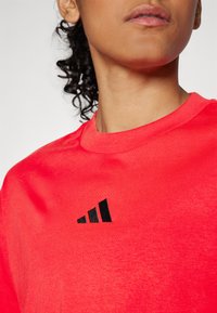 Raudonas medvilninis marškinėliai su apvalia iškirpte ir trumpomis rankovėmis, priekyje su juodu Adidas logotipu, tekstūruotas audinio paviršius.
