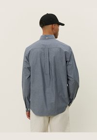 Striped button-up shirt in blue and white, featuring a back pleat, long sleeves, and a rounded hem. Accompanied by a black cap.
