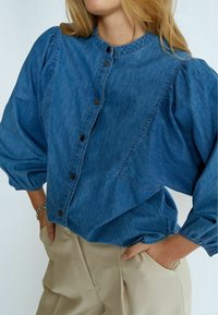 Modrá džínová halenka s kulatým límečkem, nafouknutými rukávy a zapínáním na knoflíky vpředu, s jemným prošíváním a kapsou na prsou.