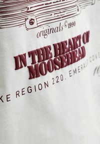 Κείμενο σε λευκό ύφασμα γράφει "In the heart of Moosehead," με μικρότερο κείμενο πάνω και κάτω, συμπεριλαμβανομένων των "originals © 1990" και των λεπτομερειών τοποθεσίας.