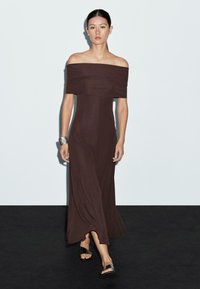 Robe maxi marron à épaules dénudées avec un bodice ajusté et une jupe fluide. Texture lisse et silhouette longue, assortie à des sandales à glissière noires.