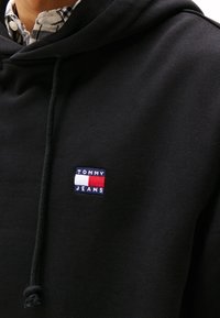 Černá mikina z bavlny s přední kapsou a šňůrkami na stažení. Na hrudi je umístěna barevná nášivka s nápisem "TOMMY JEANS".