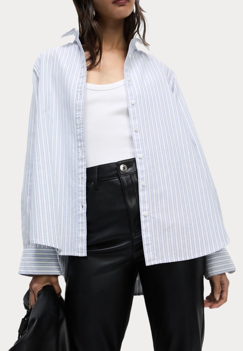 Camisa de botones de gran tamaño a rayas azul y blanca con cuello extendido, mangas largas y dobladillo ligeramente Curvado, combinada con pantalones de cuero negros.