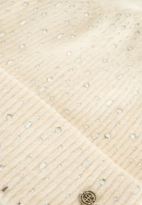 Tessuto in maglia beige con texture rigata, caratterizzato da decorazioni chiare e rettangolari sparse, e un accento di logo metallico.