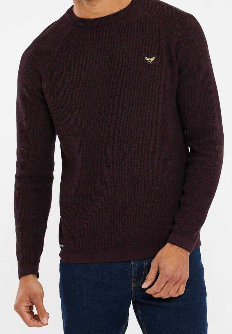 Homme portant un pull en tricot texturé marron foncé avec un petit logo jaune sur la poitrine, associé à un jean bleu.