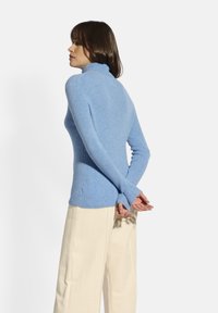 Jasnoniebieski, dopasowany sweter z golfem o prążkowanej teksturze i długich rękawach. Modelka ma na sobie kremowe spodnie z szerokimi nogawkami, z rękami złożonymi za plecami.