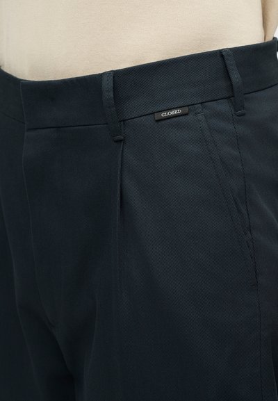 Κοντινή λήψη σκούρων ναυτικών παντελονιών με θηλιές για ζώνη και μια μικρή μαύρη ετικέτα που γράφει "CLOSED" κοντά στη μέση.