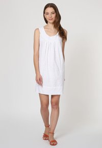 Vestido blanco sin mangas con patrón de ojal, que presenta un escote redondo y un cuerpo fruncido; combinado con sandalias naranjas de tiras.