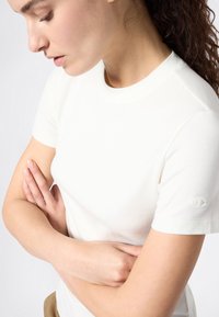 T-shirt bianco aderente realizzato in morbido cotone, con maniche corte e un piccolo logo sulla manica sinistra. Design semplice e pulito.