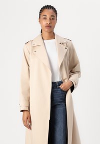Trench coat bej cu guler tip rever, mâneci lungi, butoane cu clips și buzunare laterale, purtat peste o cămașă albă și blugi negri.
