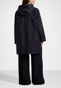 Μακρύ μαύρο αδιάβροχο παλτό με κουκούλα, που διαθέτει ίσια γραμμή, μαλακή υφή και μινιμαλιστικό σχέδιο. Φοριέται πάνω από φαρδιά μαύρα παντελόνια.